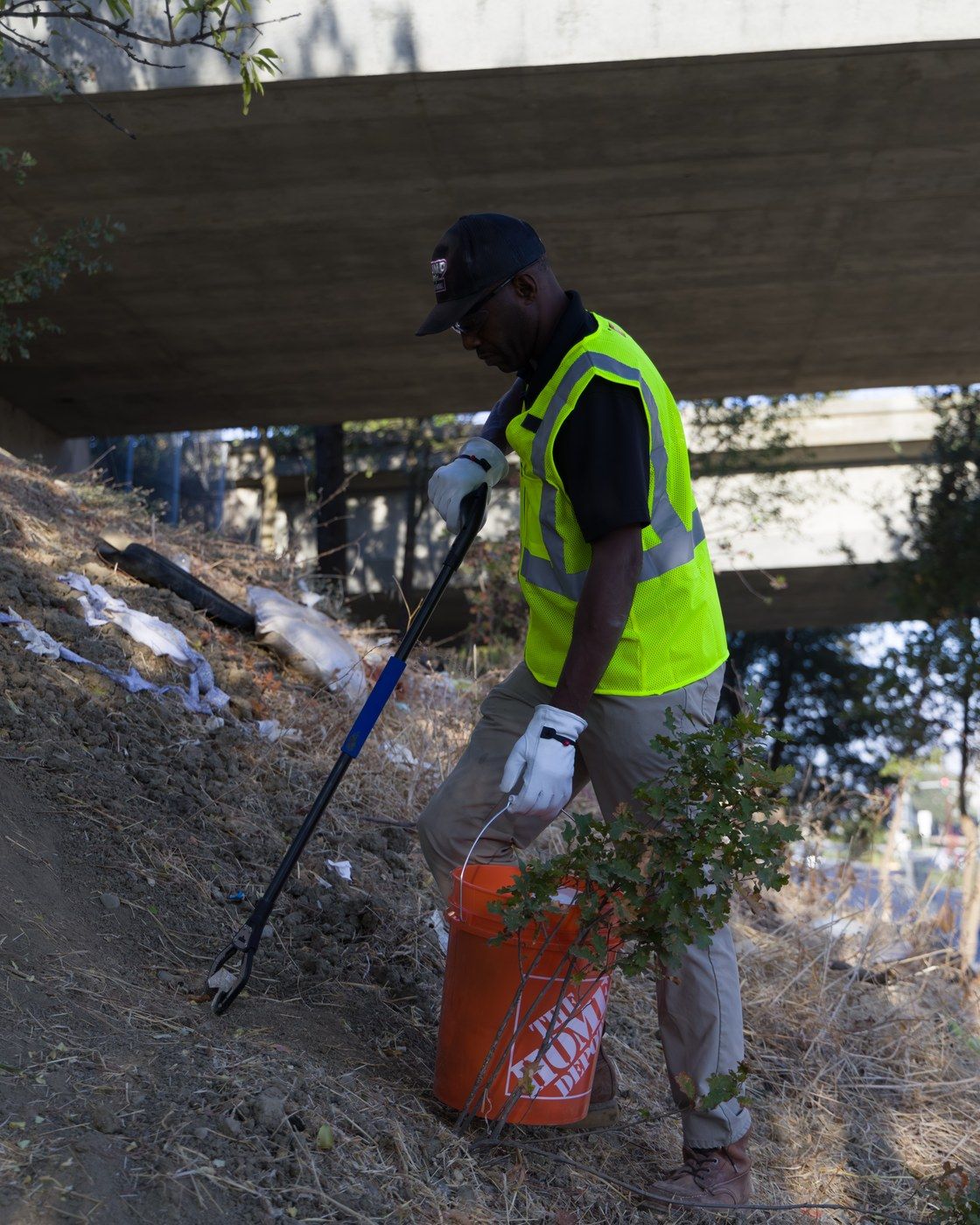 Dump Ninja crew member in a high-visibility vest picking litter under an overpass with a grabber and bucket